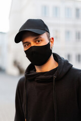 Wall Mural - portrait of handsome young hipster man model with protective medical mask and black fashion mockup cap wearing trendy hoodie in the city