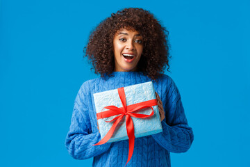 Wall Mural - Girl cant wait unwrap gifts on winter holiday season. Cute and lovely girlfriend african american female holding wrapped present and smiling, thanking standing blue background