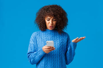 Wall Mural - Problem, messaging and people concept. Confused and displeased, tensed african-american young woman receive strange message, staring at smartphone screen clueless, shrugging, blue background