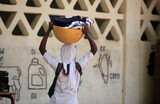 Fototapeta Most - horizontal  photpgraphy: closeup of a skinny African girl in white clothes and head wrap, carrying a full yellow bowl on her head, by the white wall- , outdoors on a sunny day, in the Gambia, Africa