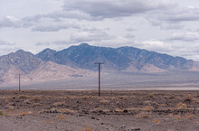 High Desert, Nevada, USA - May 17, 2011: Wide Landscape Under Gray Cloudscape With Brown Electrical Poles On Dirt With Brown Dried Shrubs. Dark Mountain Range As Backdrop.