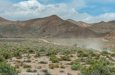 High Desert, Nevada, USA - May 17, 2011: I pickup truck and 3 SUVs drive on dirt road spewing dust in the air with mountain as backdrop under blue cloudscape. Green shrub in front.