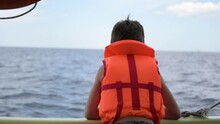 Little Child In Orange Safe Lifejacket During Summer Leisure Activity In Sea On Boat Board With Other Vessel Sailing Near