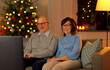 © Syda Productions - holidays, leisure and people concept - happy smiling senior couple watching tv at home in evening over christmas tree lights on background