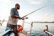 © fotofabrika - Young african american man standing with fishing rod on a sailboat fishing in open sea on sunset