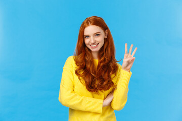 Gorgeous young redhead girl 20s in yellow sweater, smiling carefree making order, reservetion for three people, showing number third with fingers, standing blue background