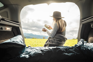 Girl resting in her car. Woman hiker, hiking backpacker traveler camper in plaid, relaxing, drinking hot tea on top of mountain. Road trip. Health care, authenticity, sense of balance calmness.