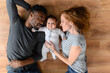 Happy mixed-race international family of three lying on the floor, happy black husband, caucasian wife and their biracial baby girl looking at the camera, top view
