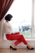 © Andriy Medvediuk - portrait of young European businesswoman with notebook is sitting by the window. Concept of planning. girl in white shirt and red pants.