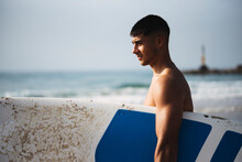 Side View Of Young Man Holding His Surfboard On The Beach.