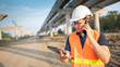 © zephyr_p - Asian worker man or male civil engineer with protective safety helmet and reflective vest using smartphone and digital tablet for project planning and checking schedule at construction site.