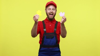 Wall Mural - Happy smiling worker man connecting two pieces of puzzles, solving hard task, integration, wearing blue uniform, red T-shirt and visor cap. Indoor studio shot isolated over yellow background.