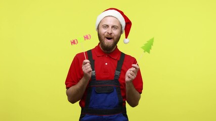 Wall Mural - Happy handyman celebrating new year eve, holding in hands Christmas party props, wearing blue overalls, red T-shirt and santa claus hat. Indoor studio shot isolated over yellow background.