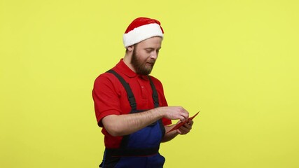 Wall Mural - Side view of touched worker holding red envelope and reading letter from mysterious stranger, wearing blue overalls and santa claus hat. Indoor studio shot isolated over yellow background.