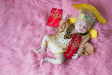 A Newborn Baby Wearing A Chinese Emperor's Costume Gold Colour Lying On A Pink Blanket. Translation On Red Envelope Is Good Luck
