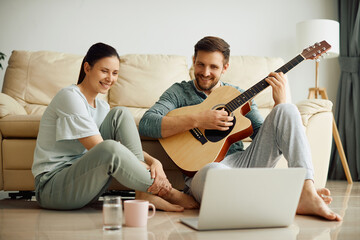 Wall Mural - Happy couple uses laptop while playing acoustic guitar at home.