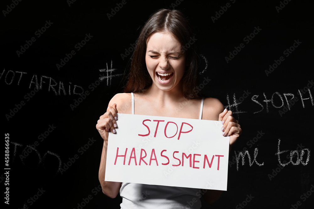 Stressed young woman holding paper with text STOP HARASSMENT on dark background