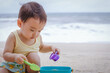 © TimmyTimTim - Asian boy child wear yellow vest playing sand with his bucket while sitting on the beach in front of sea. Phuket, Thailand. Family Holiday, summer trip concept.
