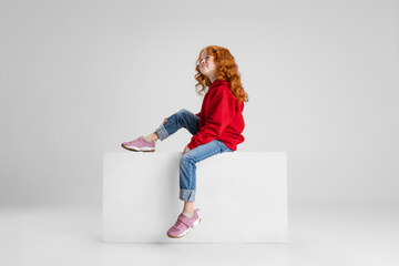 Portrait of little Caucasian red-haired curly girl dreamingly looking upward isolated over gray studio background. Side view