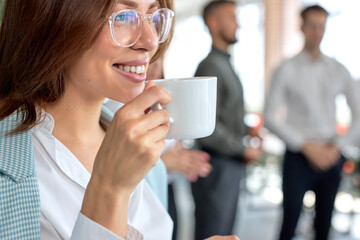 Wall Mural - Close-up Portrait Of Young Good-looking Business Manager Lady Drinking Coffee In Office, Having Rest, Taking A Break, Smiling And Looking At Side While Male Colleagues Standing In The Background