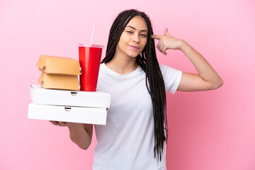 Poster - Teenager girl with braids holding pizzas and burgers over isolated pink background having doubts and thinking