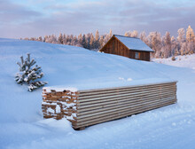 Piled Timber And Small Wooden House In Winter Snow