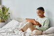 © Seventyfour - Full length portrait of smiling African-American teenager using tablet while sitting on bed in cozy room, copy space