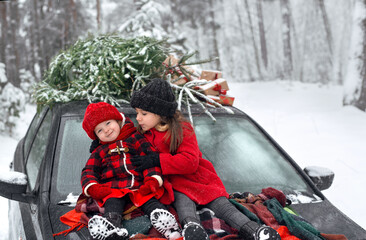 Two sisters in a checkered red and black coat, knitted wool mittens and hats are sitting on the hood of a car, next to a Christmas tree and gifts.Children in the forest on Christmas and New Year's Eve