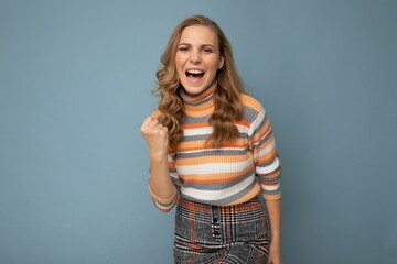 Portrait of happy positive young attractive blonde curly woman with sincere emotions wearing trendy striped pullover isolated on blue background with copy space and celebrating winning