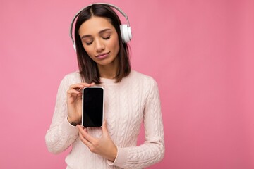 Wall Mural - Attractive positive smiling young woman wearing stylish casual outfit isolated on colourful background wall holding and showing mobile phone with empty screen for cutout wearing white bluetooth