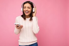 Photo Of Beautiful Joyful Smiling Young Woman Wearing Stylish Casual Clothes Isolated Over Background Wall Holding And Using Mobile Phone Wearing White Bluetooth Headphones Listening To Music And
