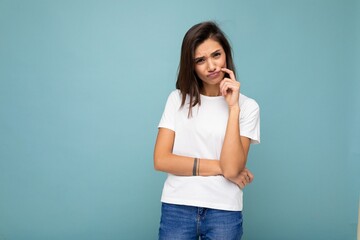 Portrait of sad thinking young pretty nice winsome brunette woman with sincere emotions wearing casual white t-shirt for mockup isolated over blue background with copy space