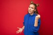 © Ivan Traimak - Photo of emotional agressive young beautiful blonde woman with two horns with sincere emotions wearing hipster bright blue hoodie isolated over red background with free space and showing fist
