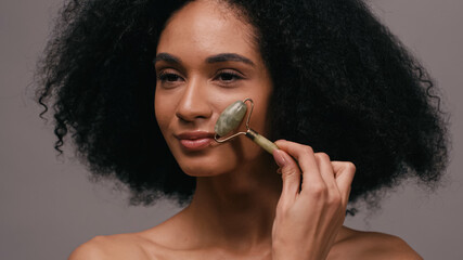 pretty african american woman with bare shoulders using jade roller isolated on grey