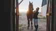 a young woman stands upright next to a horse she is holding by the halter in front of the entrance to the barn illuminated by natural light. High-quality photo