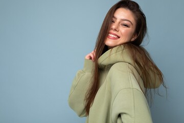 Wall Mural - Portrait of young beautiful smiling girl in stylish hipster green hoodie. Sexy carefree woman posing near blue wall. Positive model having fun