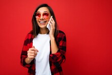 Beautiful Surprised Happy Young Brunette Woman Wearing Stylish Red Shirt White T-shirt And Red Sunglasses Isolated Over Red Background Talking On Mobile Phone Looking At Camera