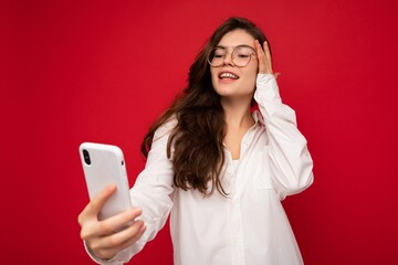 Attractive charming young smiling happy woman holding and using mobile phone taking selfie wearing stylish clothes isolated over wall background