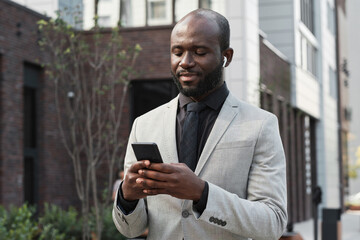 Horizontal medium portrait of joyful African American man with beard on face wearing earbuds standing outdoors choosing playlist to listen to on his smartphone