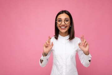 Portrait of young positive happy attractive brunette woman with sincere emotions wearing casual white shirt and optical glasses isolated over pink background with copy space and holding fingers