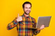 © deagreez - Photo of charming pretty young man wear plaid shirt smiling typing modern device showing thumb up isolated yellow color background