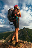 Fototapeta  - A male traveler in a red hat with a backpack is talking on the phone against the background of mountains. A man in hiking clothes talks on a smartphone at sunset in the mountains. Phone conversation