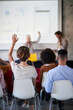 © luckybusiness - A young woman is rising her hand to ask a question while following a presentation in the office