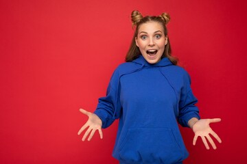 Wall Mural - Young happy positive attractive emotional joyful blonde woman with two horns with sincere emotions wearing stylish bright blue hoodie isolated on red background with copy space