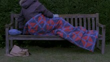 An Older Man Wearing Winter Clothing, Getting Into A Sleeping Bag, Preparing To Lie Down On A Wooden Park Bench At Twilight, With Some Belongings Nearby.
