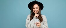 Panoramic Photo Of Attractive Young Smiling Woman Wearing Black Hat And Grey Sweater Holding Smartphone Looking At Camera Showing Shhh Gesture Isolated On Background