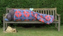 An Unidentifiable Person Wearing Winter Clothing, Having Just Got Into A Sleeping Bag, Lying Down During The Day On A Wooden Park Bench, With Some Belongings Nearby.