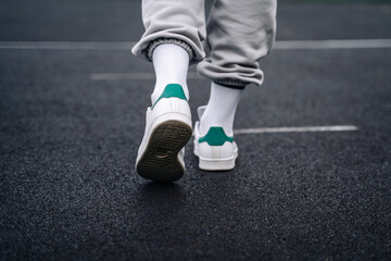 Women's legs in sweatpants, white socks and white sneakers with green elements against the background of the track with markings in the stadium.