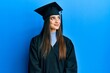 Beautiful brunette young woman wearing graduation cap and ceremony robe smiling looking to the side and staring away thinking.