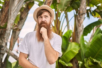 Poster - Caucasian man with long beard outdoors on a sunny day wearing summer hat serious face thinking about question with hand on chin, thoughtful about confusing idea